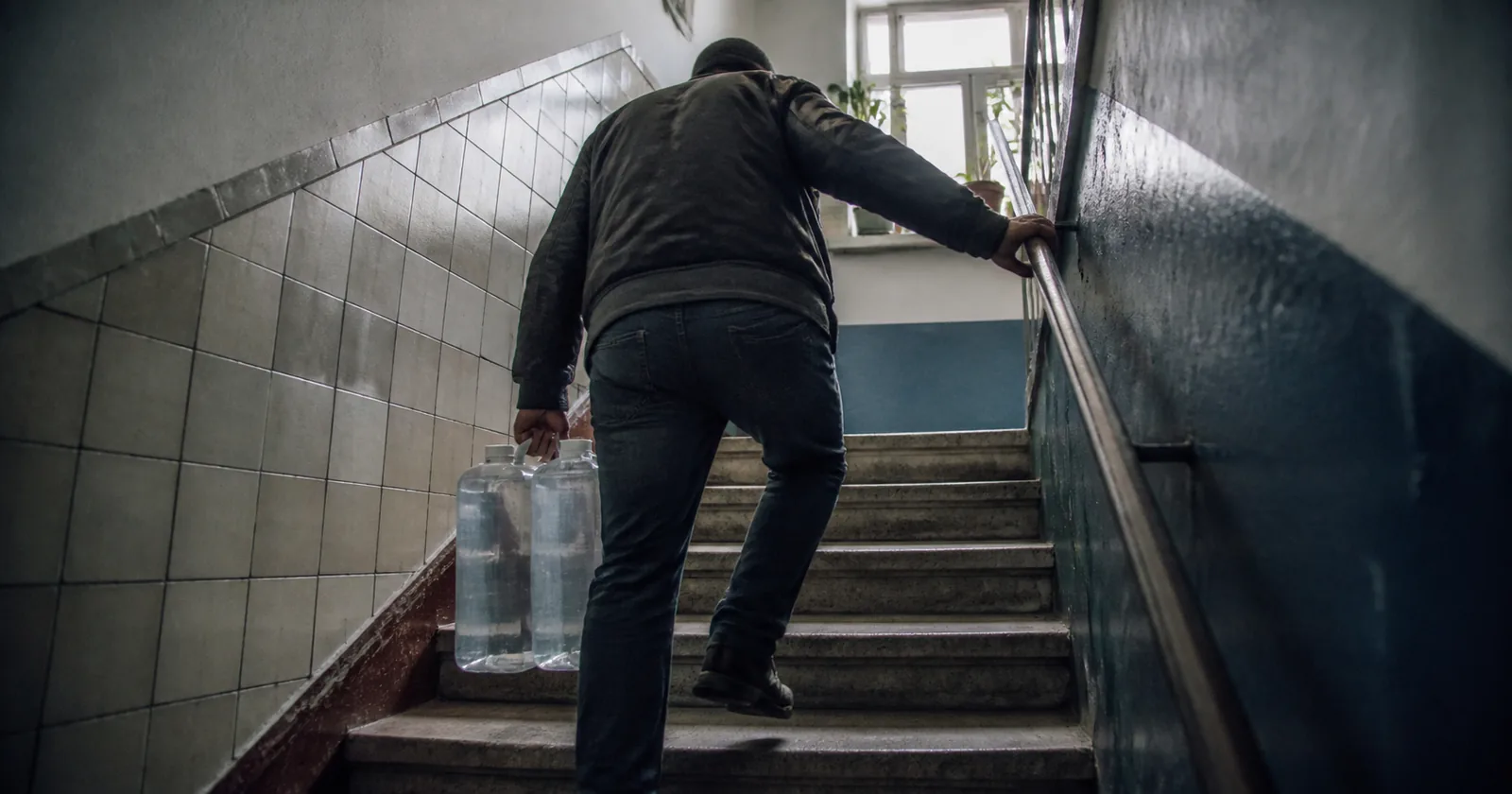 A person carrying two water containers up a narrow apartment stairwell.