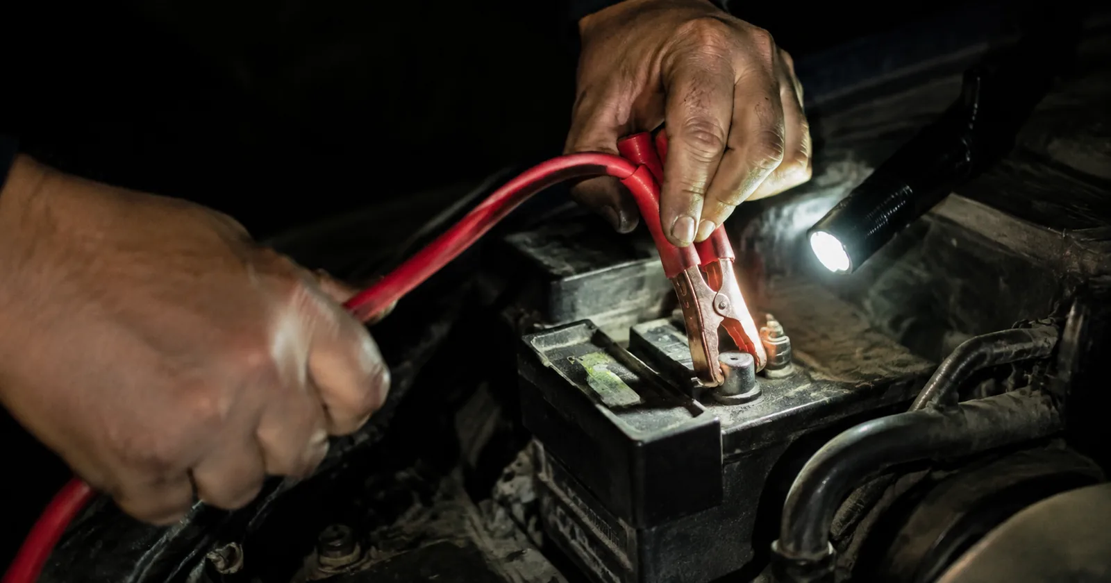 Hands clamping a jumper cable to a battery terminal at night, lit by a torch held under one arm.