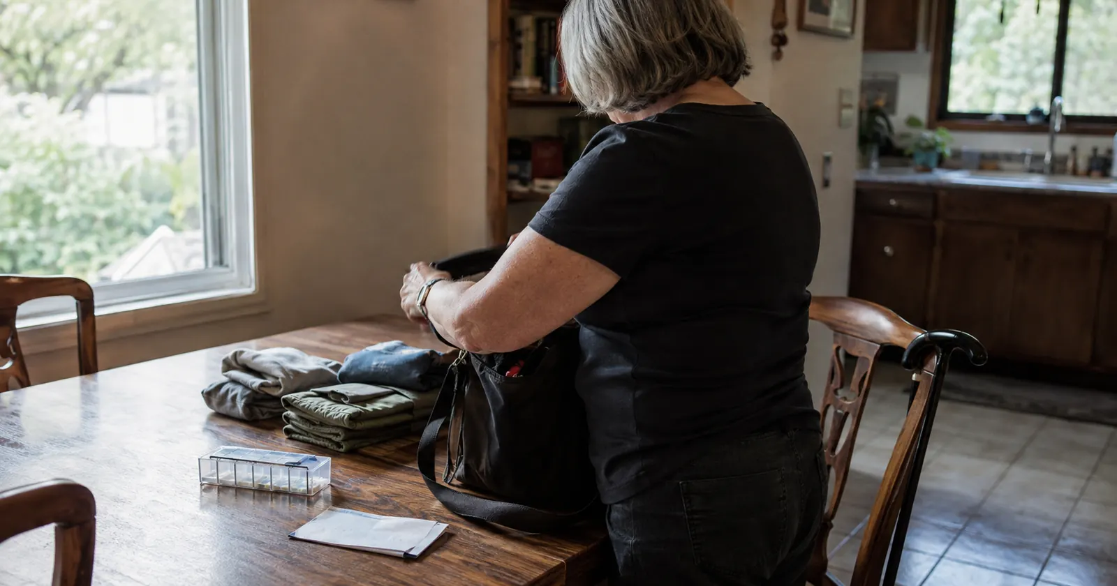 An older woman standing at a table, packing a small bag with a walking stick hooked over the chair beside her.