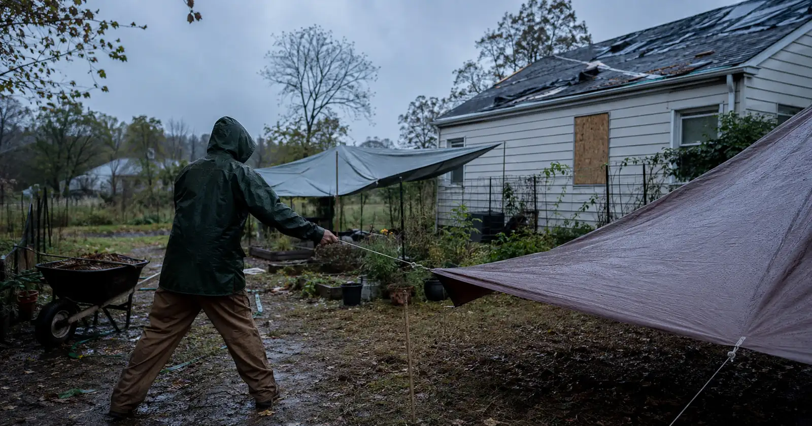 A person pulling a guy line taut on a tarp pitched beside a damaged house at dusk.