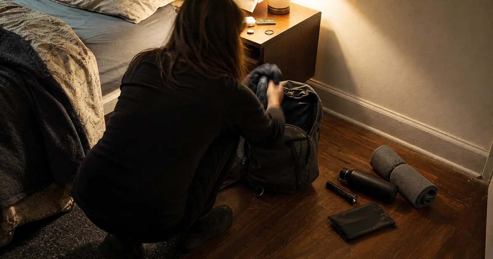 A person crouched by a bed, loading a backpack laid open on the floor in the evening.