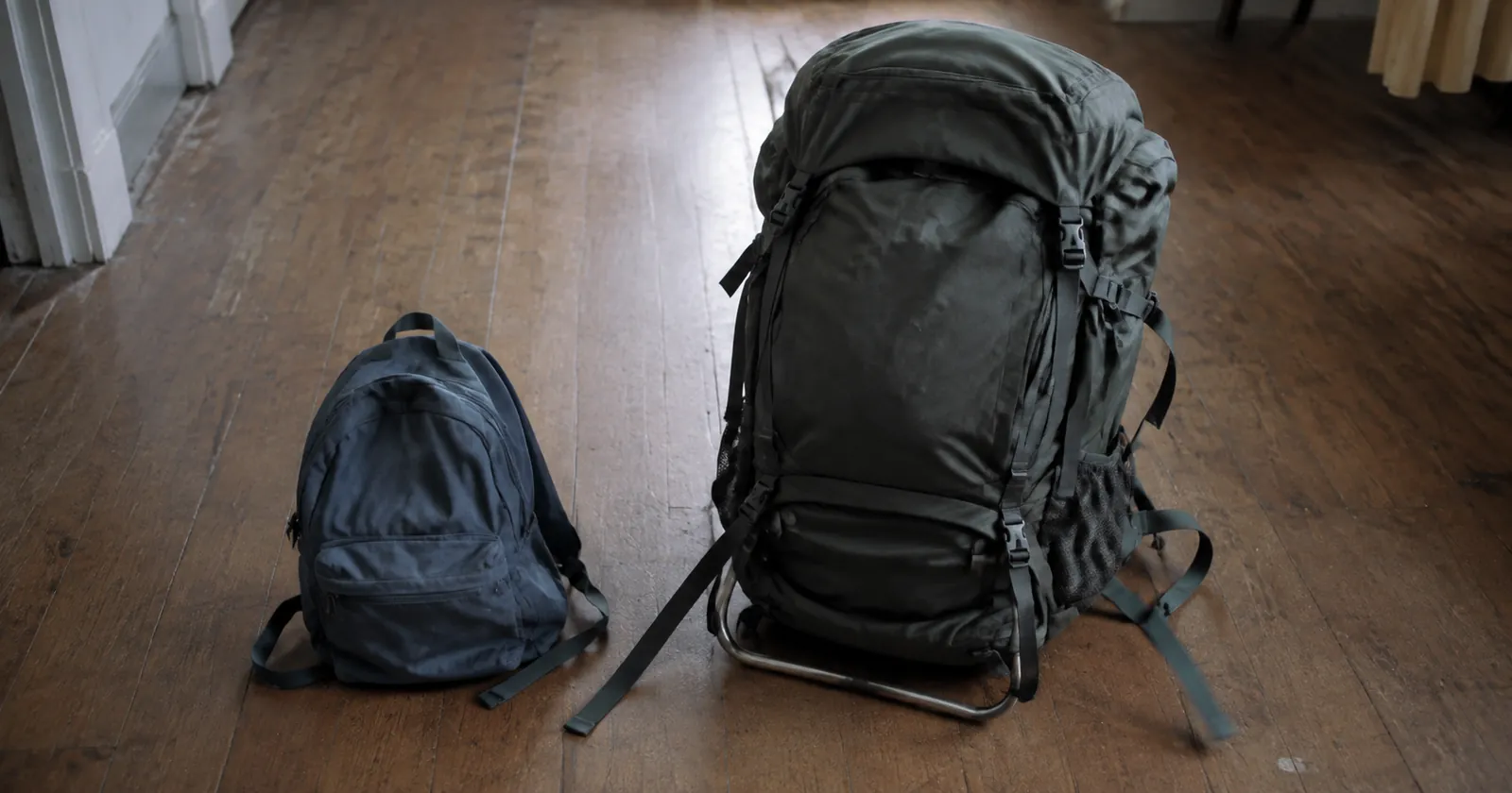 Two backpacks of different sizes side by side on a wooden floor, one small and soft, one large with a frame.